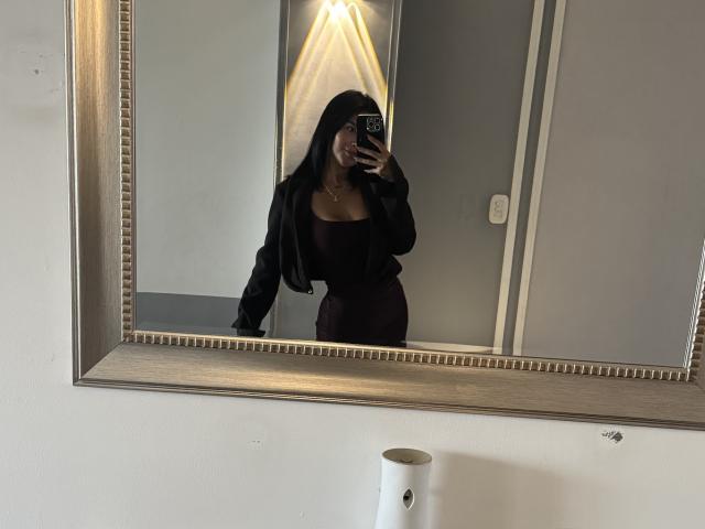 MajoJones with long black hair, wearing a black jacket and tight maroon dress, taking a mirror selfie, showing her ass and tits, in a room with a large mirror.