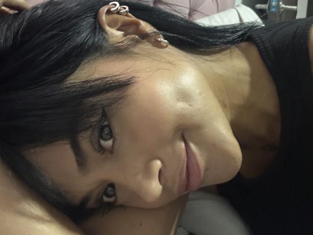 MajoJones with black hair, brown eyes, and subtle makeup, lying down, looking at the camera with a slight smile, wearing a black top, and small ear piercings.