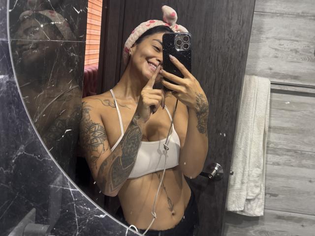 MajoJones with tan skin, tattoos, pink beanie, white sports bra showing small tits, smiling, pointing finger, holding phone, mirror selfie, black marble, white towel, wood texture.