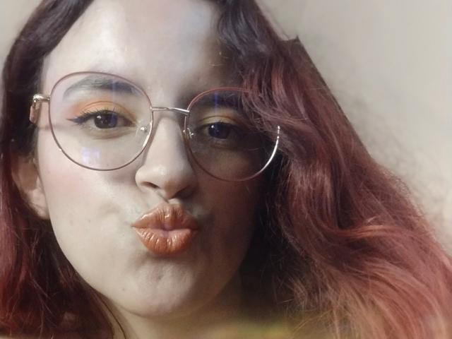 Close-up of LaiaJonnes with light brown skin, wearing round glasses with thin metal frames. She has wavy, shoulder-length hair that is reddish-brown with darker roots. She is puckering her lips, making a kissy face, with her lips slightly curled. Her eyes are highlighted with orange eyeshadow and she has subtle black eyeliner. The lighting is soft, creating slight shadows on her face. Her expression is playful. The image is focused on her face, with no visible background details. The overall composition is centered.