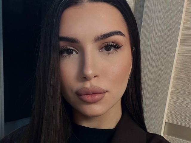 Close-up of KeenaJaminet with long, straight dark brown hair. She has full, slightly parted lips and dark, well-defined eyebrows. Her large, almond-shaped eyes have long, thick eyelashes. Her skin is smooth and fair. She wears a black top with a collar. The lighting is soft, highlighting her facial features. The texture of the background is slightly rough, contrasting with her smooth skin.