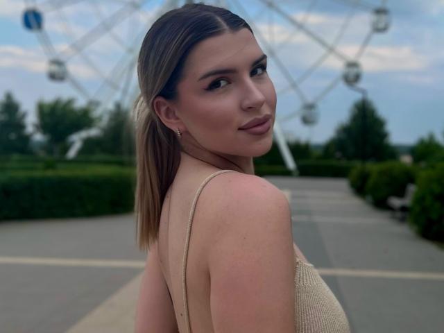 KeenaJaminet is in the foreground, facing sideways but looking back at the camera. She has fair skin, dark brown straight hair, and is wearing a beige knit top with thin straps. Her eyebrows are dark and well-defined, and she has subtle makeup with a matte brown lipstick. Her right shoulder is slightly turned, revealing a small red mark. The background has a blurred Ferris wheel and greenery. The image uses soft lighting and a shallow depth of field.
