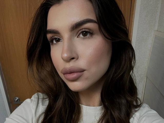 KeenaJaminet has smooth, light skin with subtle blush, full pink lips, and wavy brown hair. Her brown eyes are slightly upturned with long lashes. She's wearing a white top.