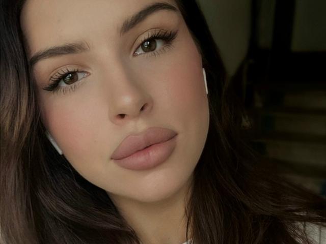 Close-up of KeenaJaminet's face. She has light skin, brown eyes, and full, pink lips. Her dark brown hair is slightly wavy and falls to the sides of her face. She's wearing white earphones. KeenaJaminet's eyebrows are thick and well-groomed, and she has long, dark eyelashes. The lighting is soft, highlighting her cheekbones and subtle blush. The overall composition is centered, focusing on her facial features. The image has a realistic, natural style.