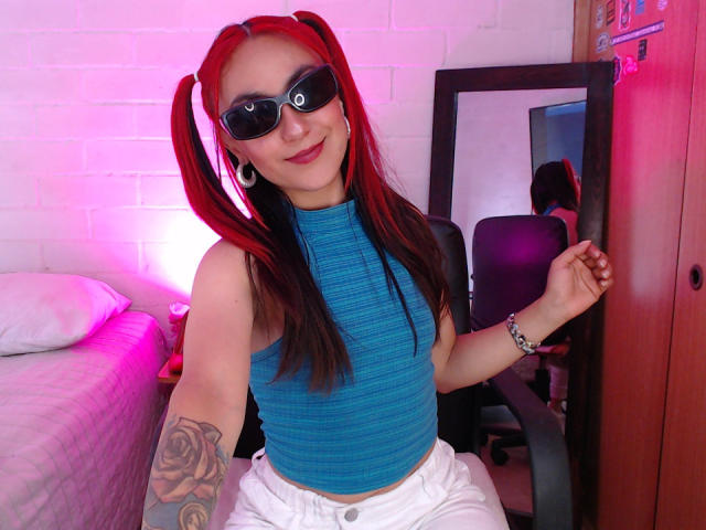 AmyStreetwise69 with red pigtails, wearing black sunglasses, blue ribbed crop top, white pants, and a rose tattoo on her left arm, sitting in a chair, smiling.