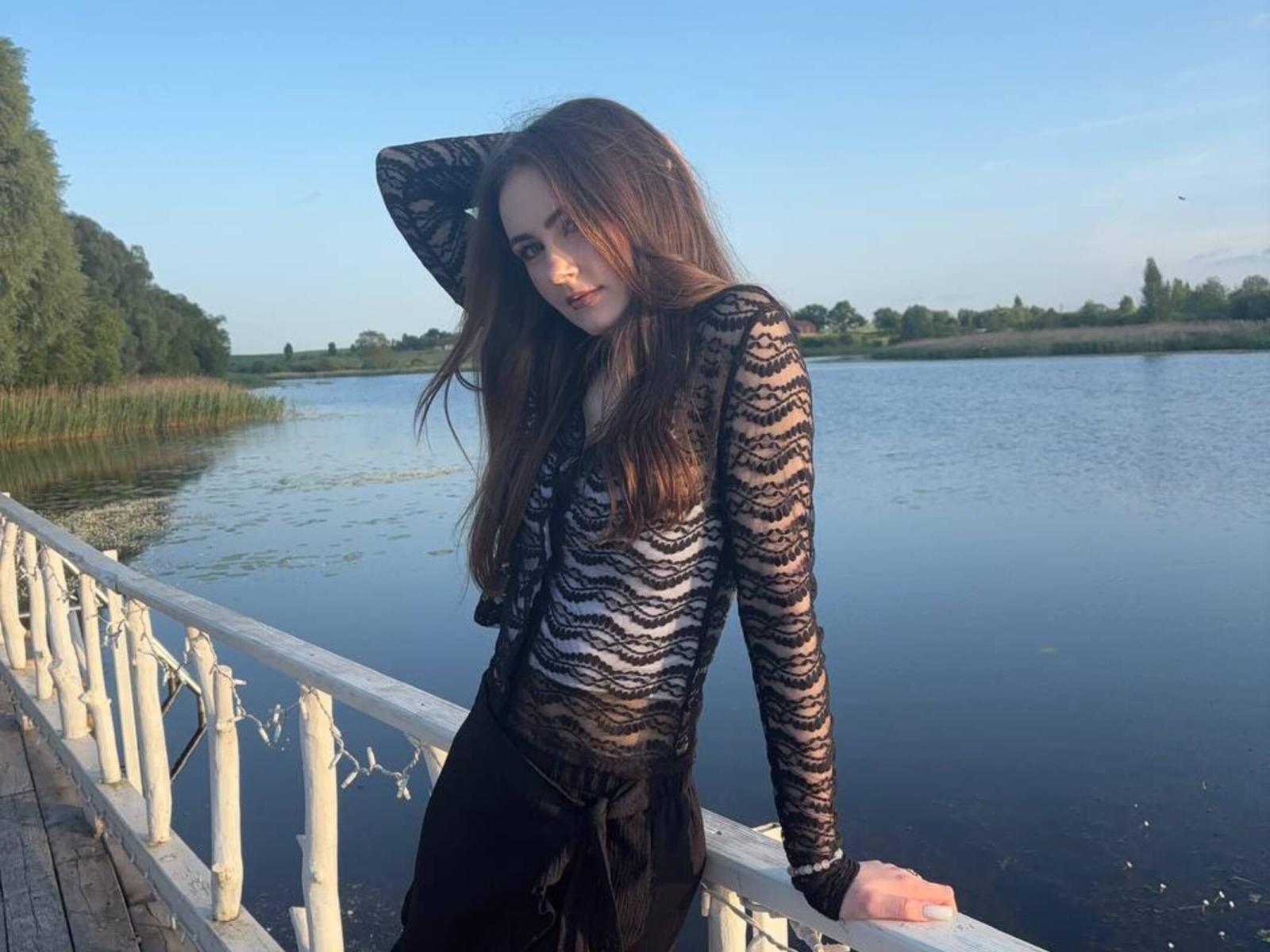 OfeliaMiessner with long brown hair, wearing a black lace top, leaning on a white railing by a calm river, her right hand in her hair, under a clear blue sky.