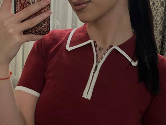 Close-up of KeenaJaminet, a woman with medium brown skin, showing her upper chest and face. She's holding a phone with a patterned case in her right hand. She's wearing a red, short-sleeved, zip-up shirt with white trim. Her black hair is tied back. She has a small silver necklace and a red bracelet on her left wrist. The background is blurred, with white and patterned elements. The image focuses on her shirt and face, with a slight smile. The photo is casual and slightly angled.