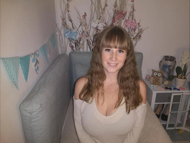 JojoKiss-hot with wavy brown hair, smiling, wearing a beige off-shoulder sweater, showing her tits, sitting on a gray couch, with a cute owl ornament on a white shelf.