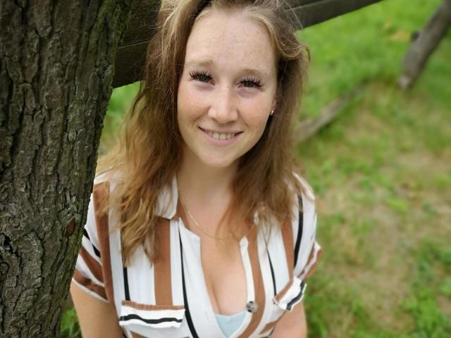JojoKiss-hot with wavy brown hair, smiling, wearing a striped shirt, standing against a tree, showing small tits and a light blue bra, with green grass in the background.