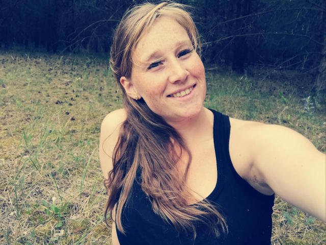 JojoKiss-hot, a chubby white woman with long blonde hair, smiling, wearing a black tank top, showing a slight armpit hair, in an outdoor grassy setting.