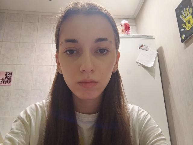 KeraPopham has long brown hair, light skin, and a neutral expression. She's wearing a white shirt, and her small, unremarkable tits are slightly visible. Bathroom setting.