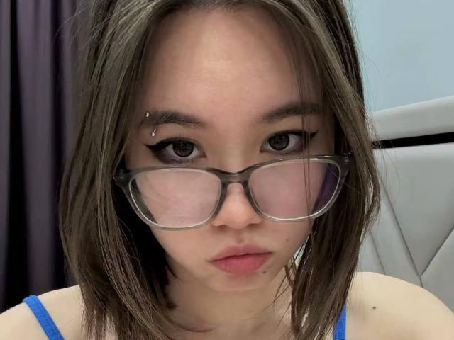 EmelineLattig with brown hair, glasses, and a small piercing on her right eyebrow, wearing a blue top, has a neutral expression and slightly parted pink lips.