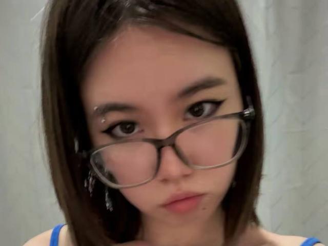 EmelineLattig has brown eyes, glasses, and short brown hair with a slight side part. She's wearing a blue top, looking serious with a small pimple on her chin.