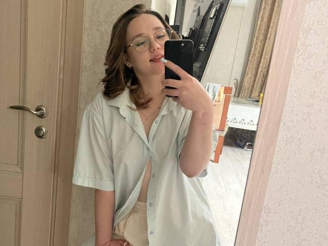 AnitraMaccoll, avec des lunettes, une chemise vert clair, les seins apparents, un soutien-gorge beige, et un téléphone dans un selfie pris devant un miroir, montrant une bouche légèrement ouverte et une expression détendue..