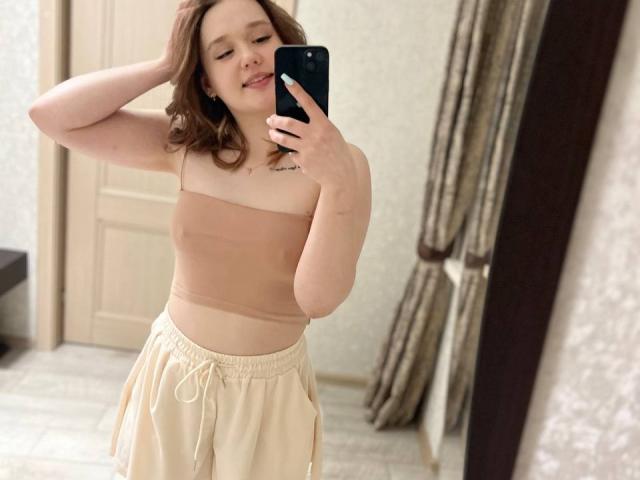 AnitraMaccoll mit mittelgroßer Brust, trägt ein beiges, trägerloses Oberteil und beige Hosen, macht ein Selfie vor einem Spiegel mit ihrem Handy, braunes Haar, lächelt, Hand im Haar, steht drinnen..