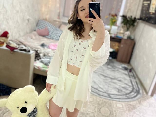 AnitraMaccoll, con un busto de tamaño medio, sosteniendo un oso de peluche blanco, vistiendo una camiseta corta blanca con estampado floral, pantalones cortos blancos y una camisa blanca abierta, tomando una selfie..