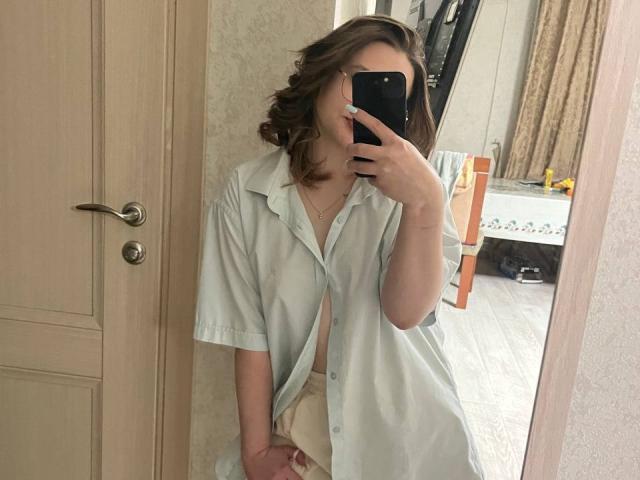 AnitraMaccoll, con i capelli castano ondulati, indossa una camicia bianca larga e sta scattando un selfie allo specchio con il suo telefono, mostrando una borsa beige in mano, in una stanza semplice..