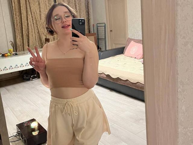 AnitraMaccoll met een bril, roze lippenstift, een bruine top en beige broek, die een vrede-teken laat zien, een spiegel-selfie, met een telefoon in de hand, in een slaapkamer, met een vloer van licht hout, een bed met een roze kussen..