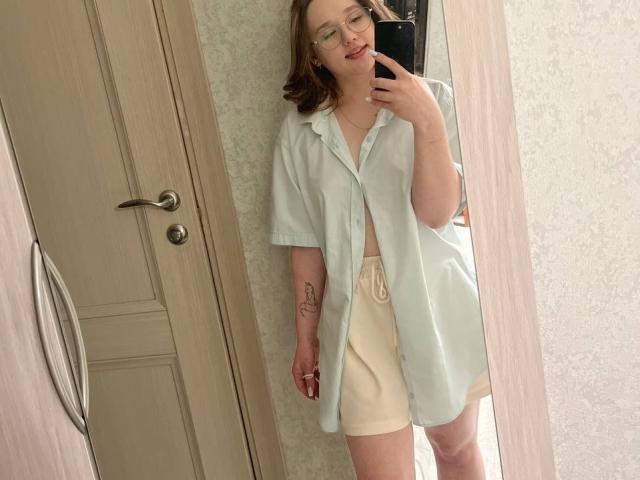 AnitraMaccoll, usando óculos, camisa verde clara, shorts bege, segurando o celular, selfie no espelho, cabelo loiro, tatuagem discreta no braço direito, porta e moldura de madeira ao fundo..