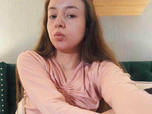 VioletaNova with long brown hair, pouty lips, wearing a pink dress, arms crossed, green studded headboard in background, taking a selfie.