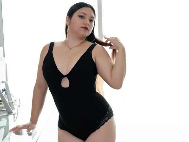 LolaDupont stands in a bright room wearing a black, deep V-cut one-piece with a small circular cutout at the chest. She has light skin, dark hair in a ponytail, and red lipstick. She holds her hair with her right hand, and her left hand rests on a glass surface. Her lace-trimmed black underwear contrasts with her smooth skin. The image is clear and well-lit.