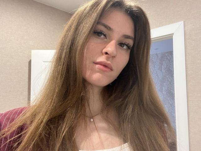 GeorgiannaWow with long brown hair, light skin, and full lips, wearing a white top and pink sweater, has a small pendant necklace, and subtle makeup with long eyelashes.