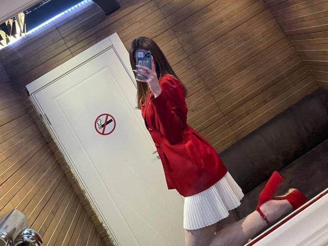 GeorgiannaWow takes a mirror selfie, kneeling on a brown sofa. She wears a red satin blazer with long sleeves and a white pleated skirt. Red high heels match her blazer. Her brown hair is down, partially covering her face as she holds a phone with a blue and white case. The door behind her has a "no smoking" sign. The wooden wall is textured in a horizontal pattern. The image focuses on GeorgiannaWow's outfit and reflection.