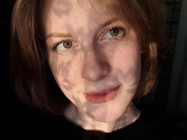 DebbyPennimpede's face, with light brown hair, green eyes, and a subtle smile, shows sun patterns on her pale skin, wearing a black shirt, in a well-lit photo.