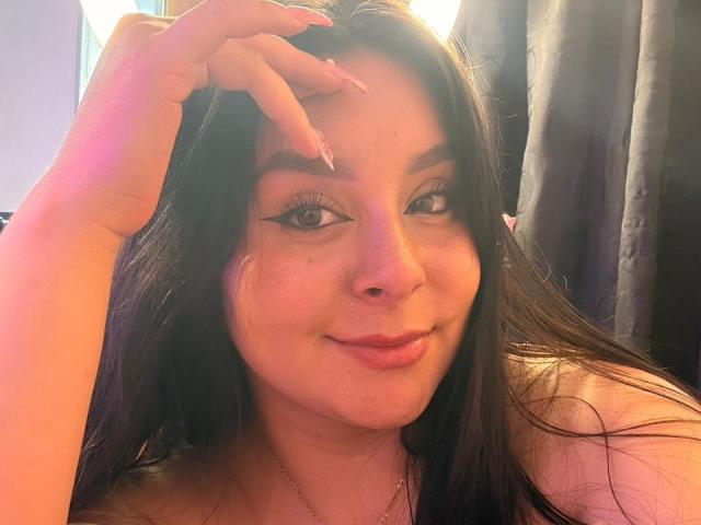 EmaJames with long dark hair, wearing eyeliner, smiling with pink lips, touching her head with manicured fingers, pinkish-orange light on her face.