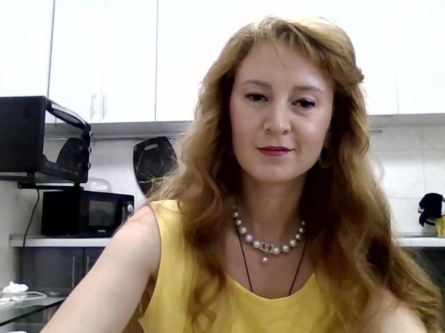 Tamariza, a young woman with wavy, light brown hair, wears a yellow sleeveless top and a pearl necklace. She has fair skin, red lipstick, and a slight smile. She's in a kitchen with white cabinets, a black microwave, and a black toaster. Her hair is loose, and her makeup is subtle. The image has a casual, selfie-like quality. The kitchen's white and black elements contrast with her yellow top.