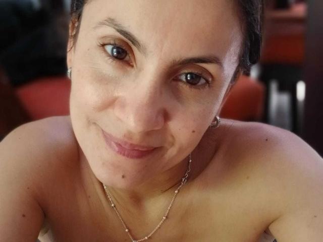 Close-up of SolaraDubois with light skin, brown eyes, and small, slightly upturned nose. She has dark brown hair pulled back and a subtle, confident smile. Her lips are slightly parted. She's wearing a thin, gold chain necklace. Her eyebrows are thin and slightly arched. She has small, dark moles on her cheek and chin. The lighting is soft, highlighting her facial features. The background is blurred, with hints of red and dark brown. The image is centered on her upper torso, showing her bare shoulders.