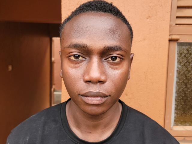 KingStone has dark brown skin, short curly black hair, brown eyes, and is wearing a black shirt with a neutral expression. His nose is slightly upturned and he's looking forward.
