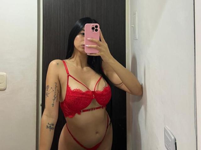 Isabella James, avec de longs cheveux noirs, un soutien-gorge et une culotte en dentelle rouge, tenant un téléphone rose, montrant des tatouages sur ses bras, debout dans un selfie devant un miroir, montrant ses seins et ses fesses..