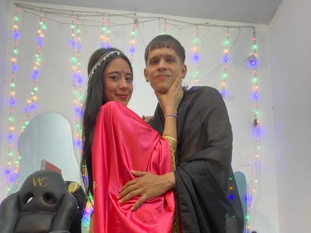 A photo of AnjaliRohan and a man standing close. AnjaliRohan has long black hair, wearing a pink traditional dress, and a white headband. The man has short brown hair, a mustache, and wears a black traditional outfit. Both have red marks on their foreheads. AnjaliRohan is being held from behind by the man. A black snowboard with "WC" in gold is on the left. Multicolored string lights hang in the background. The man's hand rests on AnjaliRohan's hip.