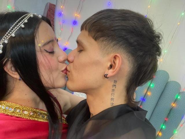 AnjaliRohan with long black hair and a pearl headband kisses a man with short brown hair and a tattoo on his neck. Both have closed eyes and are leaning in. AnjaliRohan wears a red top with gold trim, the man a black shirt. Multicolored string lights are in the background. The man has a small black earring in his left ear. The image is a close-up, capturing the kiss and their facial expressions. The man's tattoo is in Chinese characters. The composition focuses on their kiss.