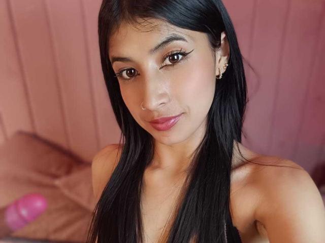 Close-up photo of NaiaSanders with long, straight black hair, medium brown skin, and a small nose ring in her right nostril. She has dark, arched eyebrows, long eyelashes, and pink lipstick. She wears small gold earrings and a black strapless top. The background is blurred, with pink and brown tones. A pink object is slightly visible on the left. Her expression is calm and slightly smiling. The image has a soft, warm light.