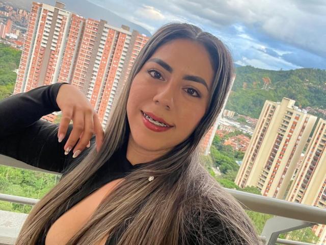 QuickHookup with long brown hair, black top, and red lipstick, smiling, showing her tits, hand on her head, standing in front of tall red and white buildings.