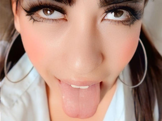 Close-up of VanellopeLm's face with brown eyes looking up, thick black eyeliner, and long eyelashes. She has dark brown eyebrows, pink blush on her cheeks, and is sticking out her pink tongue. She's wearing large silver hoop earrings and a white shirt. Skin is smooth with a slight shine. The focus is on her expressive eyes and playful tongue. The image is sharply detailed.