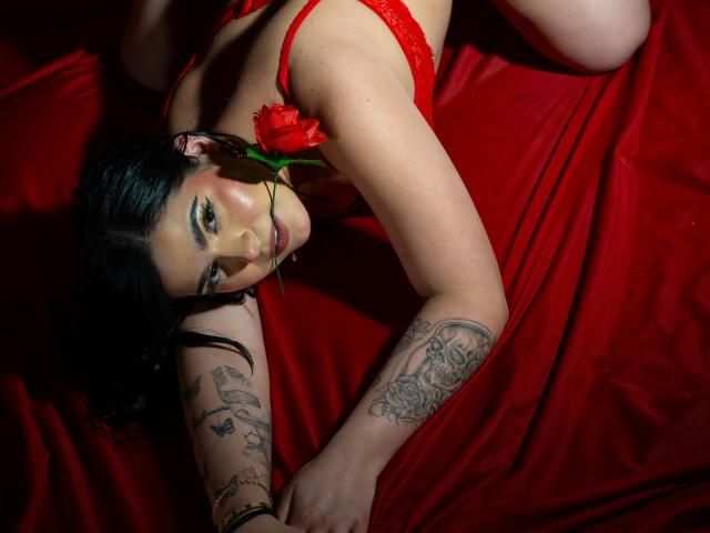 NikkyHearts is lying on red fabric, wearing red lace lingerie, with a rose in her mouth, black tattoos on her arm, and dark hair, looking at the camera.