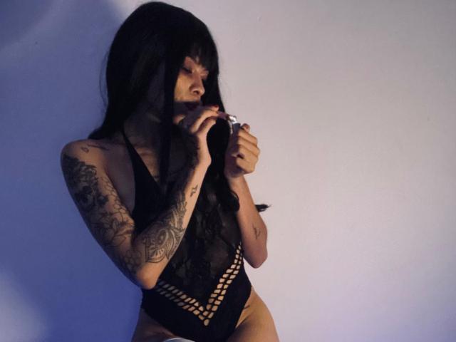 ShelsyLatin, con cabello negro largo y tatuajes en los brazos, vestida con lencería negra de encaje, sosteniendo un objeto negro cerca de la boca, mirando hacia un lado, en una habitación con poca luz..