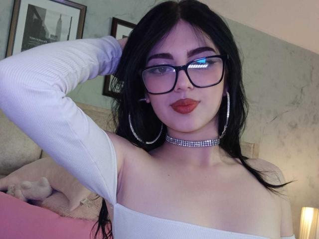 IsabellaaRossi with dark hair, glasses, red lipstick, white off-shoulder top, choker, large hoop earrings, hand in hair, smiling, light green wall, bedroom.