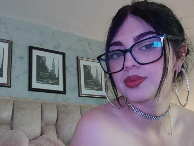 Isabella Rossi, avec des lunettes, du rouge à lèvres rouge et un collier, montrant ses épaules dénudées, les cheveux noirs relevés de manière désordonnée, portant de grandes boucles d'oreilles, souriant légèrement..