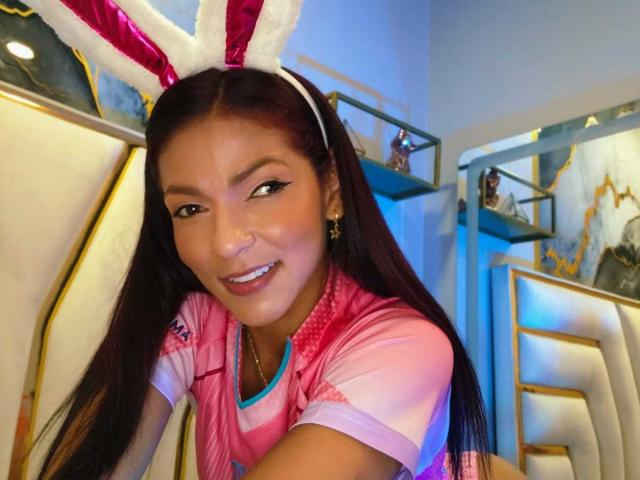 HannaPretty, an Asian woman with long black hair, wears a pink and white bunny costume with white bunny ears. She's smiling at the camera, showing white teeth. Her makeup is subtle with black eyeliner. She has gold earrings and a pink scarf around her neck. The background features a blue-lit room with shelves. The image has a soft, warm light. She has smooth, medium-toned skin. The costume has a shiny, textured fabric.