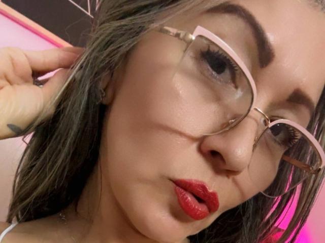 Close-up of NicoletteShea with light skin and brown hair. She wears pink-framed glasses, her right hand touching her hair. She has red lipstick and subtle eye makeup. Her eyebrows are thick and dark. The lighting is warm, highlighting her skin texture. The image has a casual, selfie-like composition. Her expression is slightly pouty. The photo focuses on her face, with a slight blur in the background. Her hair is slightly messy, adding to the casual vibe.