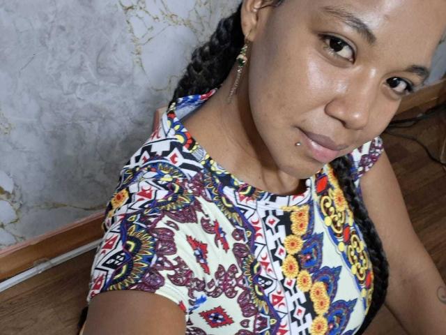NaturaleBonyy with braided hair, wearing a colorful, patterned dress, has a nose piercing and small earrings, sitting on a wooden surface, showing a subtle smile.