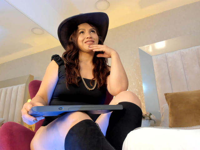 CameronViviane, a woman with light brown skin and wavy dark brown hair, sits in a red chair. She wears a black sleeveless top, a black hat, and black knee-high boots. She holds a black tablet in her right hand, resting her left hand on her chin. Her expression is slightly curious, looking upward. The room has a beige wall with a mirror and a white bed with brown pillows in the background. The image has a warm, soft light.
