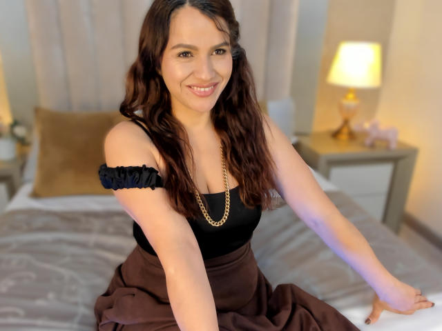 CameronViviane, a woman with long, wavy brown hair, is sitting on a bed. She has a light skin tone and is smiling at the camera. She's wearing a black off-shoulder top with a gold chain necklace and a brown skirt. Her right arm is resting on the bed while her left arm is out of frame. The background includes a blurred bed with a beige pillow and a lamp on a nightstand. The image is brightly lit with a warm, soft light. Her hair and top contrast with the brown skirt, creating a balanced composition.