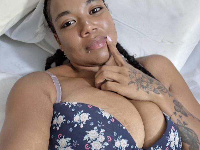 NaturaleBonyy lying on a bed, wearing a floral bra, touching her pouty lips with a finger, showing her large tits, braided hair, and tattoos on her hand.