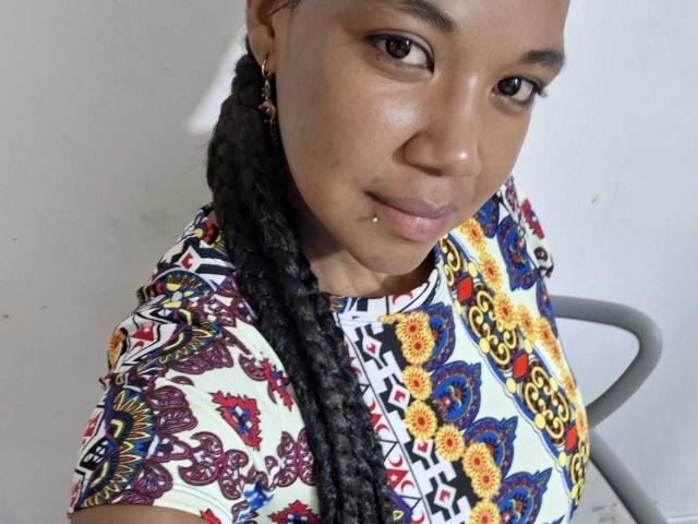 NaturaleBonyy, a young Black woman with medium brown skin, has a small lip piercing on her lower left lip. She has long, thick, twisted black hair in a single braid over her right shoulder. She wears a colorful, patterned shirt with red, yellow, blue, and white geometric and floral designs. Her eyes are slightly down, and she has a subtle, confident smile. Her right ear has a small gold hoop earring. The image is a close-up, focusing on her upper body and face.