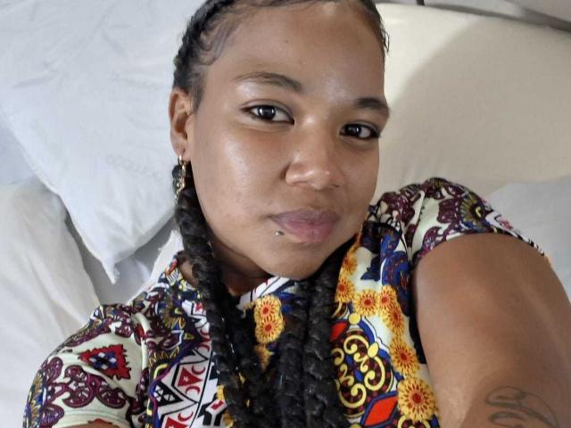 NaturaleBonyy has a medium-brown skin tone, with black, tightly braided hair. She wears a colorful, patterned shirt featuring red, yellow, and blue floral designs. She has small, dark earrings and a visible lip piercing. Her brown eyes look directly at the camera with a slight, neutral expression. A tattoo is visible on her right arm. The image is a close-up selfie.